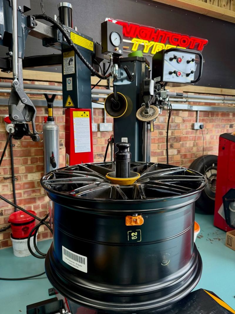A large black alloy wheel clamped on the tyre changer, the red and yellow Knightcott Tyres neon sign above it