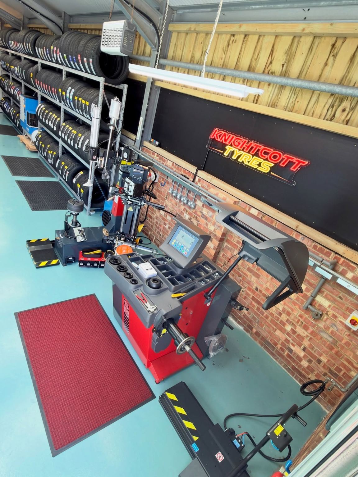 Inside the Knightcott tyre centre: racks of new tyres, a red Sice wheel balancer and a tyre changer, the Knightcott Tyres neon sign on the wall