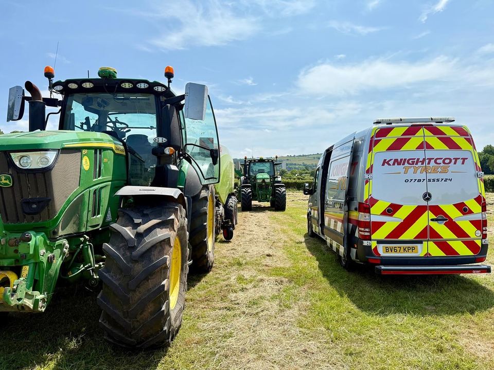 The silver Knightcott Tyres van parked beside a green John Deere tractor in a cut hayfield, a baler working behind